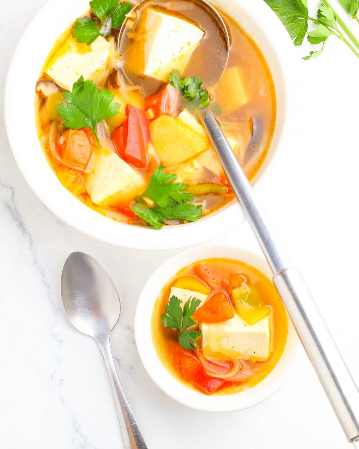 one large bowl of lemongrass thai soup with big spoon and one small bowl with a spoon beside it on a white table